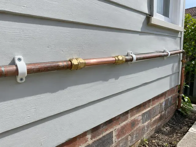 Repaired copper water pipe on an exterior weatherboard wall above a brick base in Newcastle West